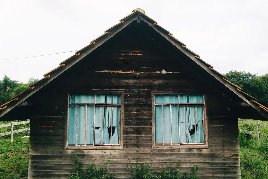 Exterior of aged abandoned residential house with shabby windows and torn curtains located on overgrown ground in countryside